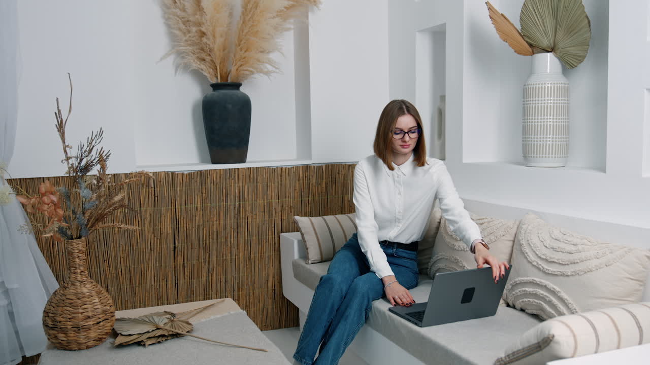 Brunette woman sitting on the sofa opens her laptop. Lady takes the gadget on her laps and starts working.
