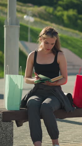 lady seated on outdoor bench with shopping bags on both sides, retrieves a book from mint green bag, opens it, and goes through its pages, background includes lamp poles, greenery
