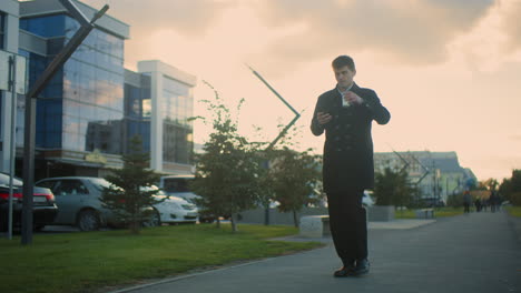 investor in black coat walking outdoors while drinking coffee and using phone in vibrant urban environment with glass buildings, greenery, parked cars, and soft sunlight