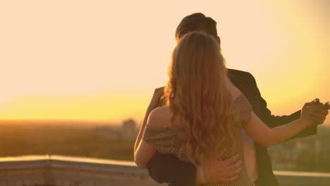 un hombre y una mujer enamorados bailan de pie en el techo de un edificio al atardecer mirándose el uno al otro.