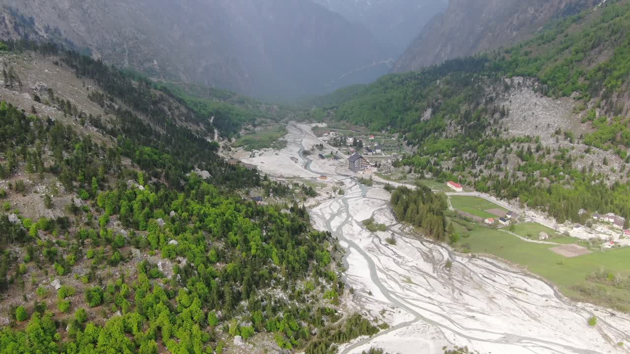 Arnavutluk'ta Alpler üzerinde uçan, karlı zirvelere sahip dağlarla çevrili bir vadide yeşil ormanı ve Valbon'da akan nehirleri gösteren drone görüntüsü.