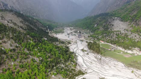 Drone-view-in-Albania-flying-in-the-alps-showing-green-forest-on-a-valley-surrounded-by-mountain-with-snowy-peaks-and-rivers-flowing-in-Valbon?