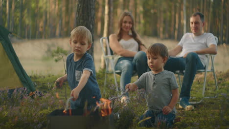 Two-boys-3-6-years-together-fry-on-fire-marshmallows-on-sticks-against-the-background-of-parents.-Family-hike-in-the-woods-with-a-tent.-Family-in-nature-on-a-hike