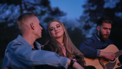young friends rest in nature at dusk. beautiful woman listen to a boyfriend while sitting together at camping party. guy playing the guitar.