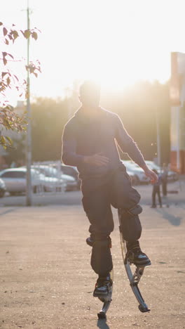 front shot of man wearing gray hoodie and dark gray pants jumping on spring stilts on paved road in residential area surrounded by houses during sunset, with serious expression