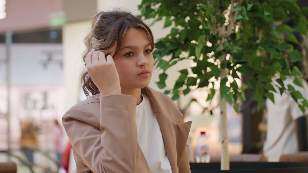 Joven pensativa, mujer blanca alisándose el pelo bajo la vegetación, chica con chaqueta informal reflexionando cerca de una planta de interior, joven ajustándose el pelo al lado de un árbol de interior en un centro comercial