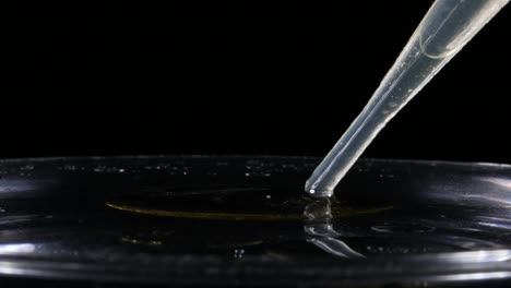 Dropper-quickly-pushing-oil-onto-surface-of-water-on-black-background,-Closeup