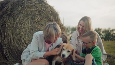 mujer y hija con niños jugando con perro con correa en el campo abierto, estrecho abrazo afectuoso, rodeados de balas de heno al atardecer, familia disfrutando del campo