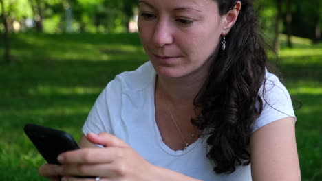 plano medio de una mujer joven sentada en el césped en el parque desplazándose por las redes sociales en su teléfono inteligente y riendo