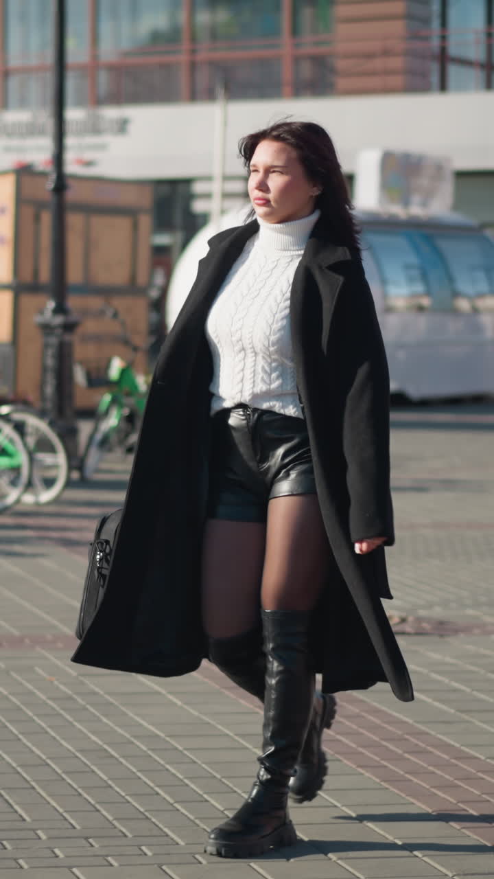 자신감 있는 여성이 비즈니스 미팅에 걸어가고, 세련된 검은 코트와 부츠를 입고, 주차 된 자전거와 함께 바쁜 도시 거리에서 그녀의 뒤를 따라 고 있습니다.