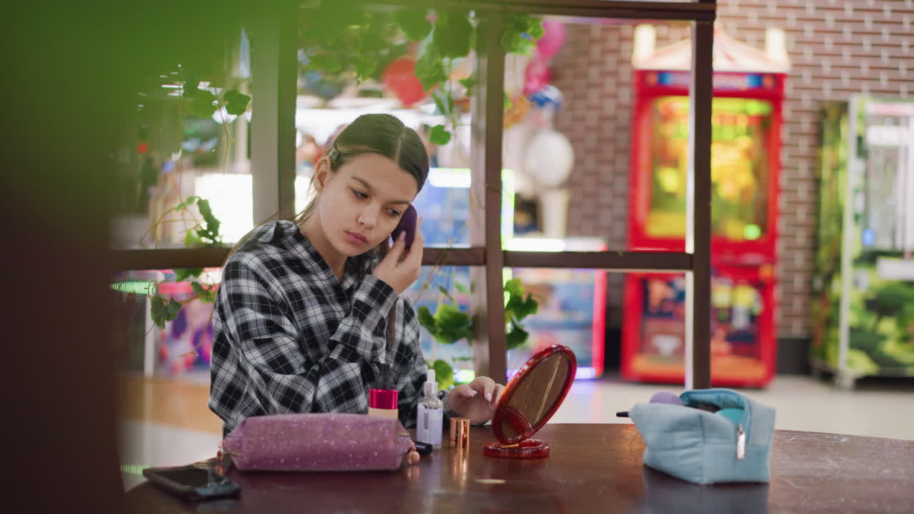 mujer joven en camisa a cuadros ajustando el maquillaje en la cara con esponja mientras mira en el espejo, productos de belleza cuidadosamente dispuestos en una mesa de madera en el espacio interior animado máquinas de arcade en el fondo