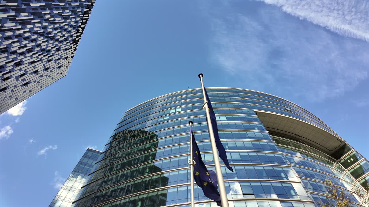 European Union flags fly beside the Lex Building in Brussels, framed by modern glass architecture and a blue sky with airplane contrail