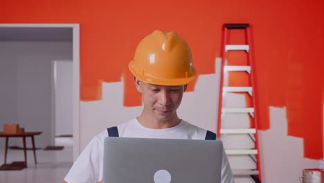 close up de un pintor asiático que usa un casco de seguridad usando una computadora portátil mientras está de pie pintando la pared en la habitación