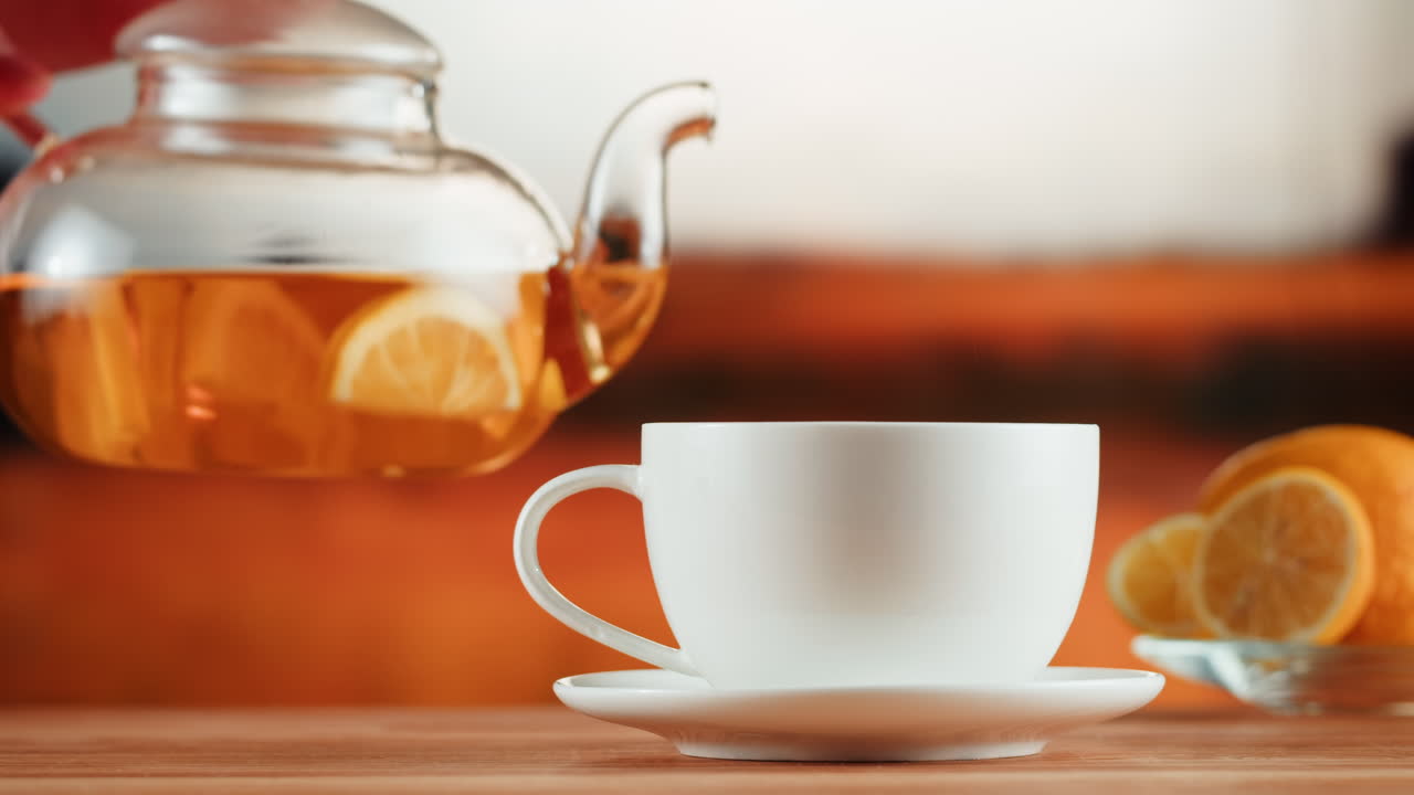 Pouring hot lemon tea from a glass teapot into a white cup