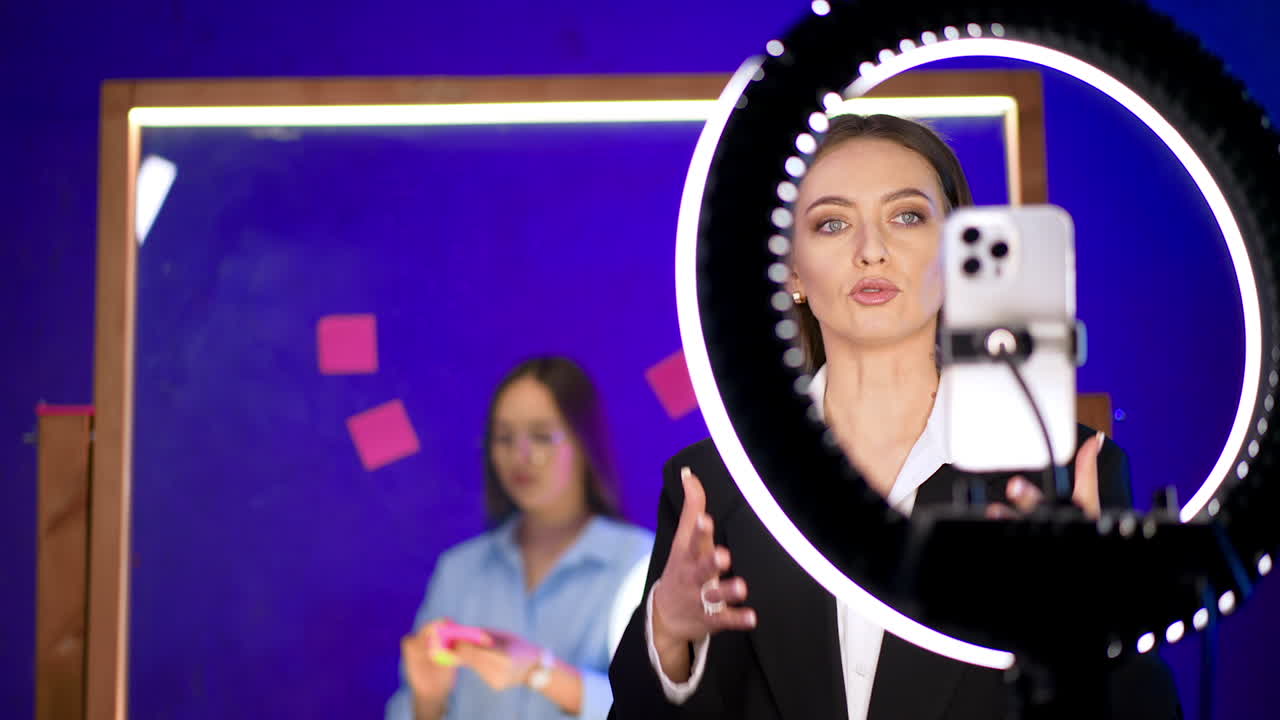 Lady talks in front of her phone and ring light. Woman records a video or streaming for blog. Girl at backdrop puts stickers on the glass wall.