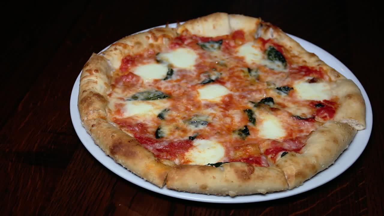 A close-up shot of a freshly baked pizza on a white plate, featuring a thick crust topped with tomato sauce, melted mozzarella, and fresh basil, held over a wooden table.