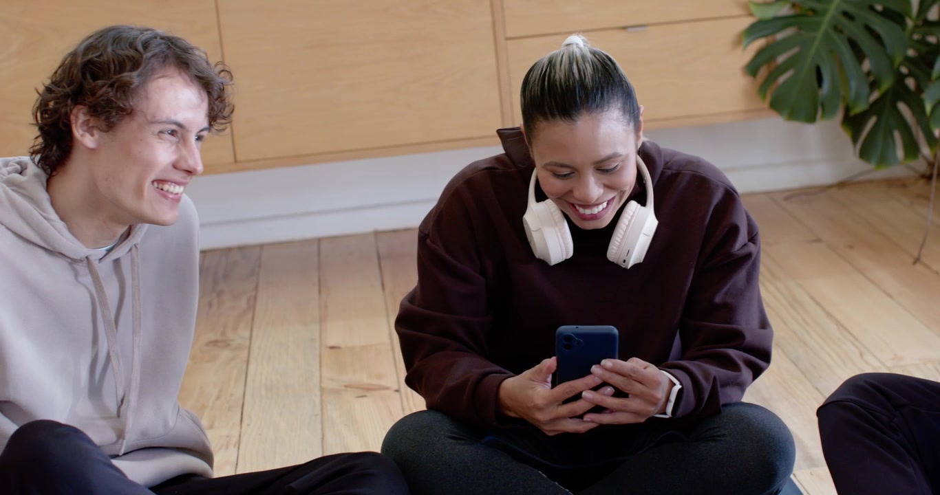 Diverse friends watching woman scrolling phone after ping laughing while leaning on wood floor