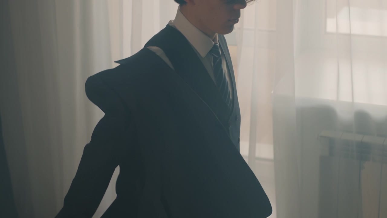 Musicista asiatico che si sistema il gemello prima di un'esibizione con luce soffusa dalla finestra, gilet e abito formali, sedia con giacca appoggiata, rituale tranquillo nel backstage, preparazione concentrata e sottile tensione catturata