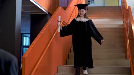 happy preschool female student in cap and gown running down the stairs, holding graduation diploma and hugging teacher