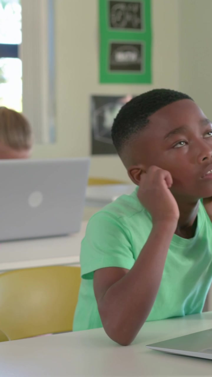 Vertical video: Camera panning boy in green lifting head rubbing face thinking in class with laptop