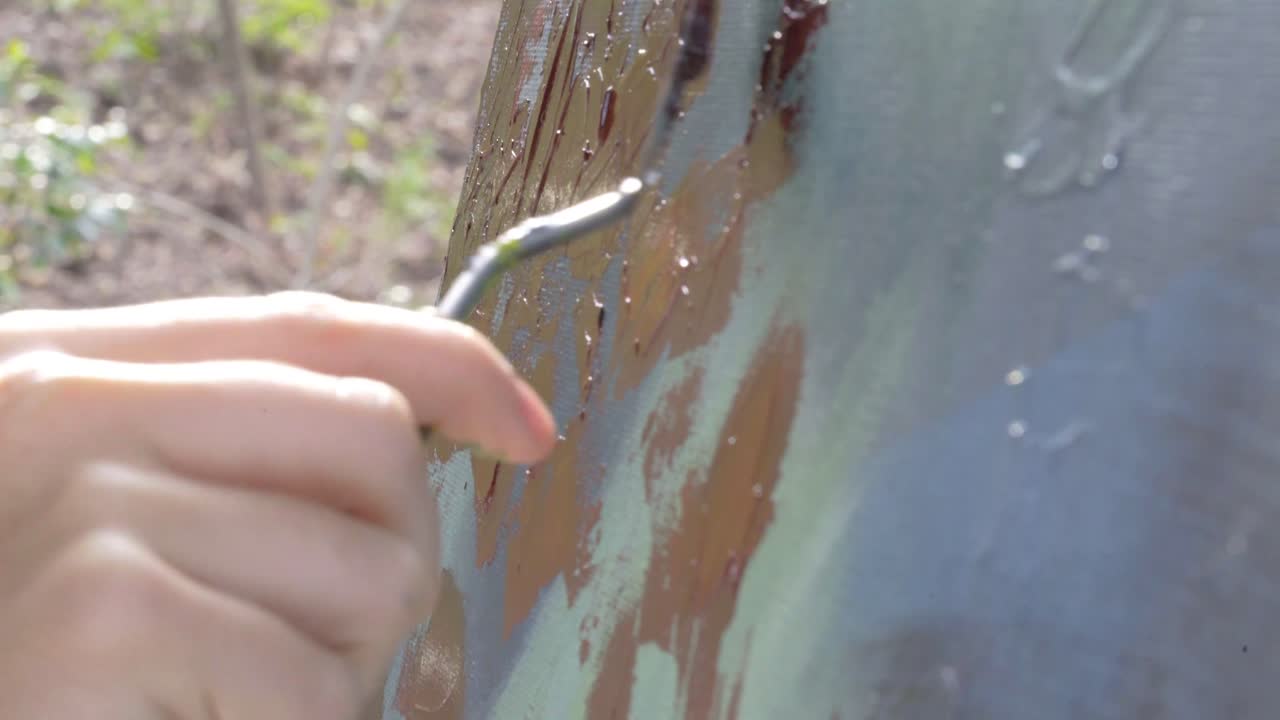 Female artist smearing red paint and creating texture on multicolored abstract painting outside during the day, camera tilt down