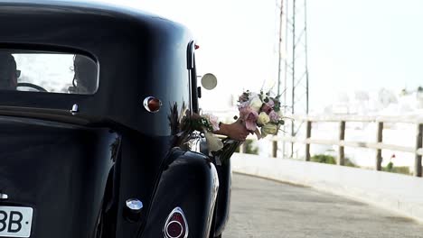 Slow-motion-backward-dolly-shot-of-the-black-vintage-wedding-car-of-the-groom-and-bride-while-the-bride-holds-a-bouquet-of-roses-out-of-the-window