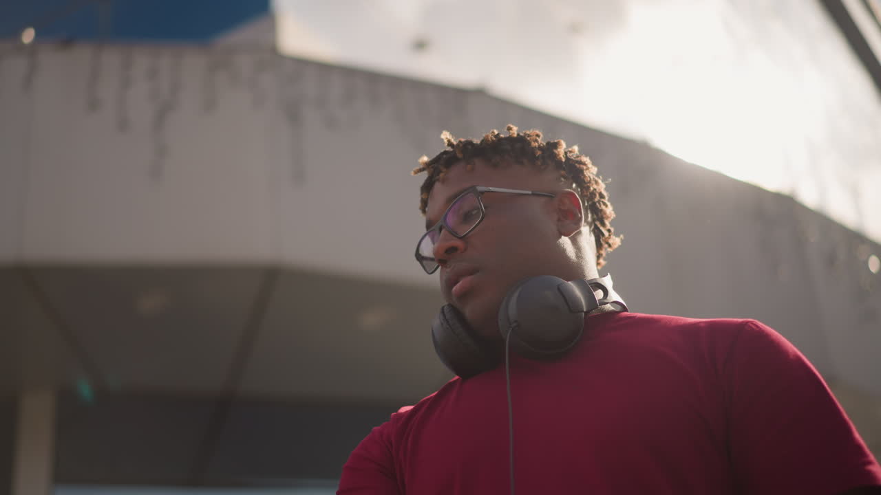 DJ di colore ripreso dal basso che mixa all'aperto in una piazza moderna, cuffie al collo, maglietta rossa, espressione concentrata, giradischi e controller visibili, sfondo architettonico illuminato dal sole, artista di strada sicuro di sé