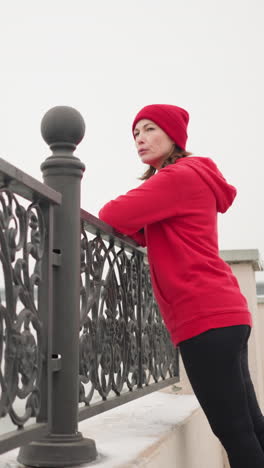 mujer con capucha roja y leggings negras descansa la mano en la valla de metal decorativa mientras mira el puente lejano con coches en movimiento, río tranquilo en el fondo, rodeado de tierra cubierta de nieve durante el invierno