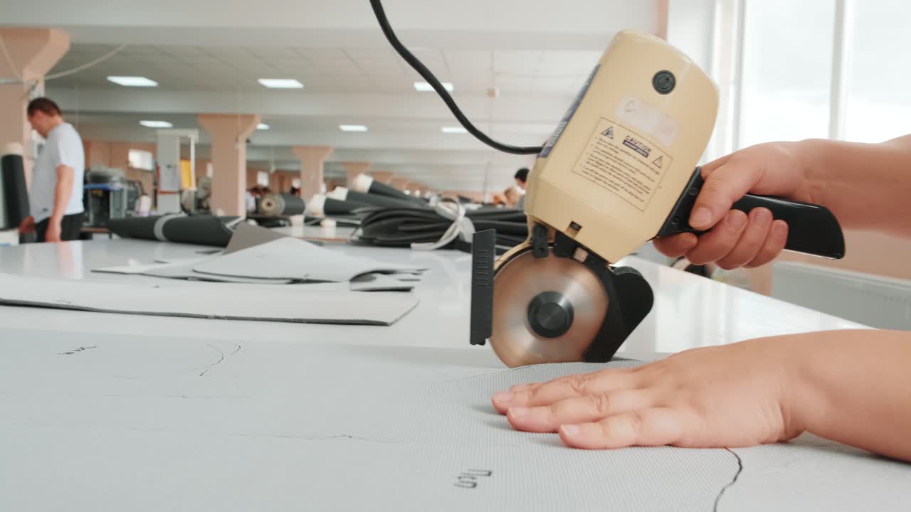 Female worker on a sewing manufacture uses electric cutting fabric machine. Sewing production line