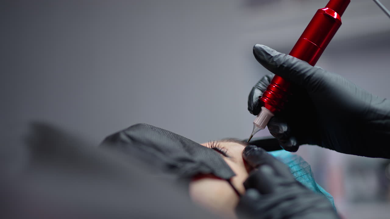 Black instrument for permanent make-up in the hand of professional cosmetologist in black gloves. Young woman is getting permanent lips make-up. Close-up