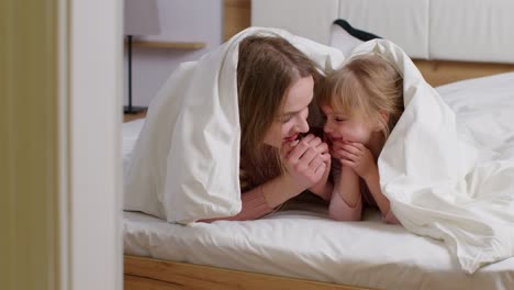 happy family of mother with daughter lying on bed under duvet blanket, telling secrets to each other