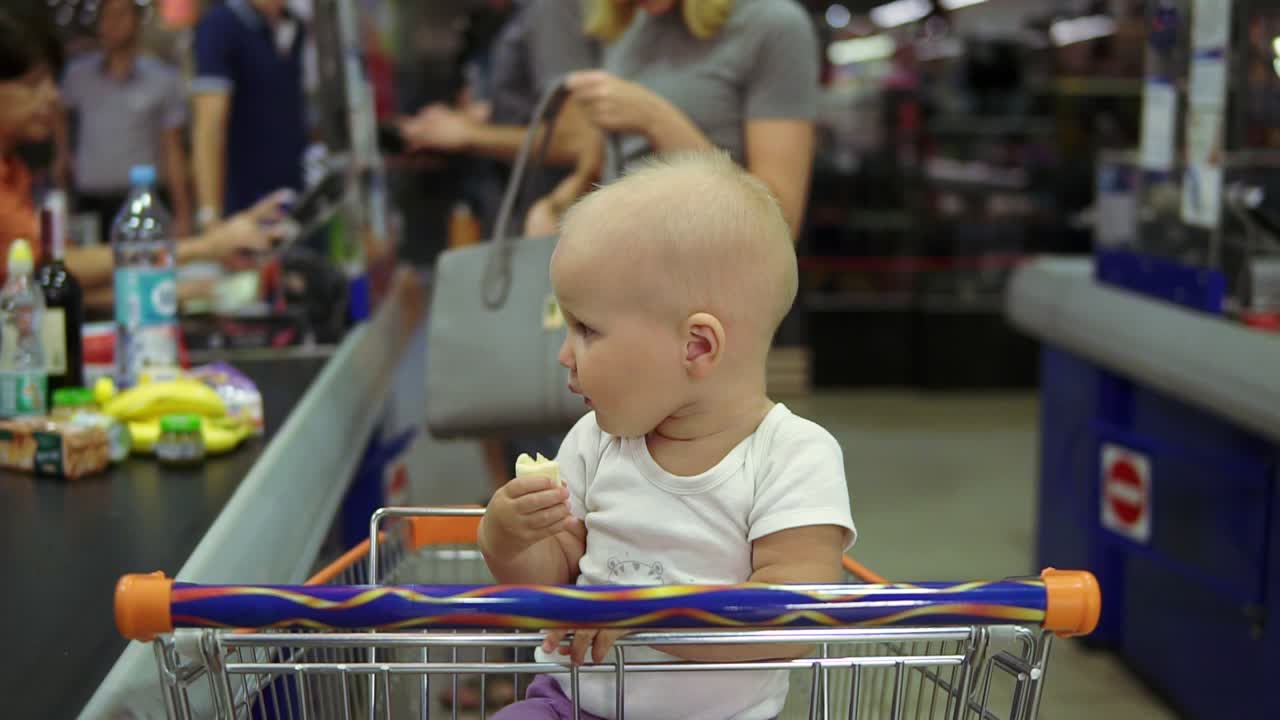 vista de cerca de un niño pequeño tomando plátano y sentado en un carrito de compras mientras su madre está sacando productos de la
