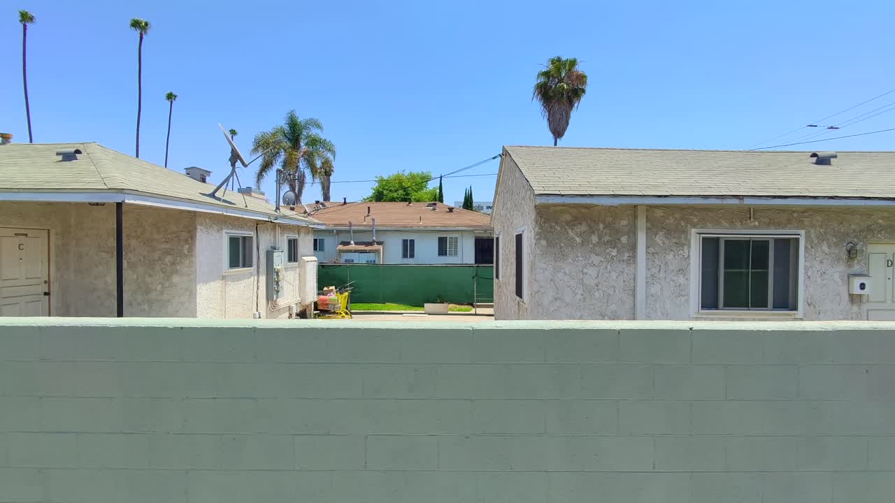 Beautiful Kill Bill like panning shot of a green wall with a quaint house like apartment building and palm trees in front of brilliant blue sky background. Drop text on the wall or in the sky easily