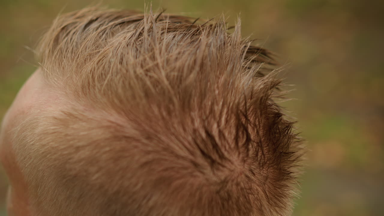 músico blanco con un mohawk corto peinándose antes de un concierto, manos dando forma a las puntas húmedas, recinto al aire libre borroso, llovizna al atardecer, ambiente de ensayo vanguardista, primer plano de la textura y el cuero cabelludo, preparación para la actuación