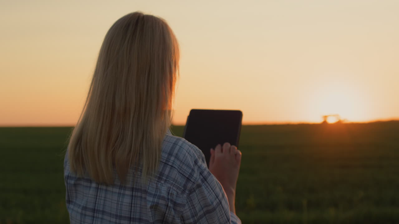 A farmer woman uses a tablet. Standing in a field where a tractor is working in the distance and the sun is setting