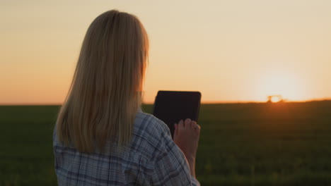 a farmer woman uses a tablet. standing in a field where a tractor is working in the distance and the sun is setting