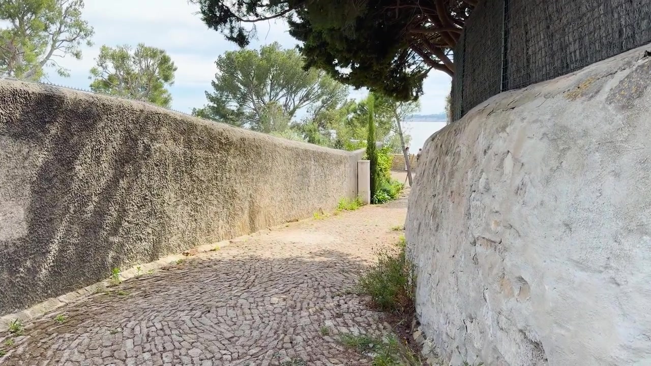Uma panorâmica lenta com câmera na mão segue um caminho de paralelepípedos curvo ladeado por muros de pedra rústicos provençais