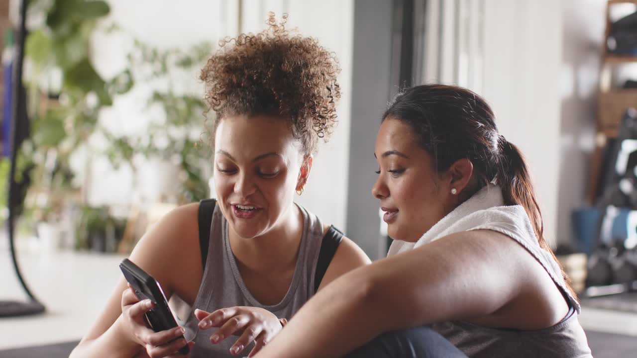 Diverse female friends sitting on gym floor after workout retrieving smartphone reviewing results