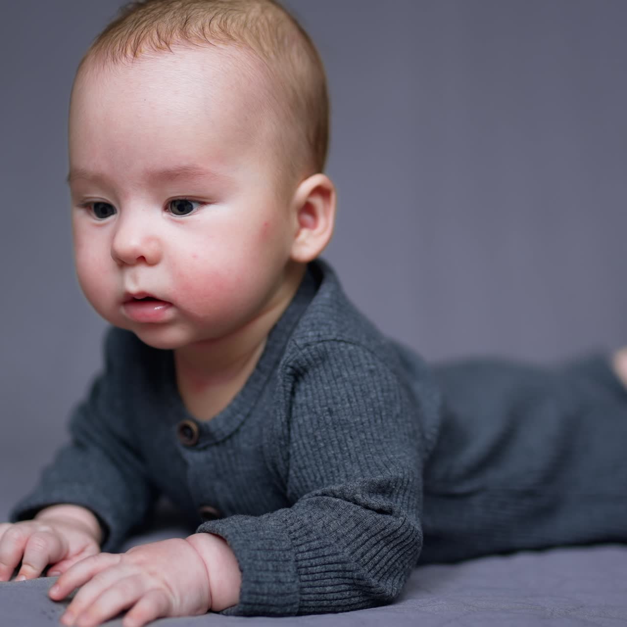 Healthy lovely kid in grey clothes lies on his belly. Adorable child looking sideways with interest. Grey blurred backdrop