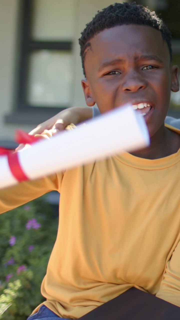 Vertical video: Boy in yellow waving diploma on porch, girl placing arm around him for graduation