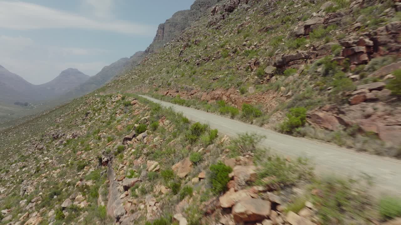 un avión no tripulado vuela rápidamente al borde de una carretera y un paisaje rocoso en el área silvestre de cederberg en sudáfrica con una vista de un vehículo todoterreno que conduce en una carretera.