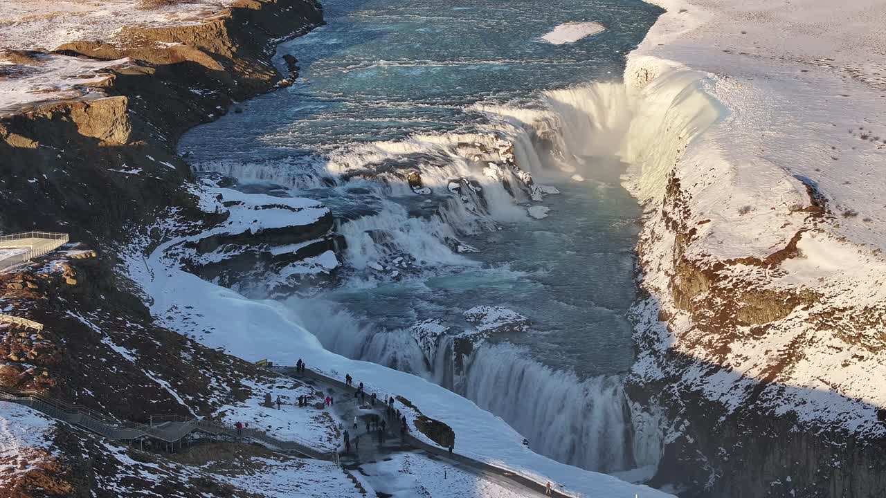 An aerial view of Gullfoss waterfall in Iceland during winter, showcasing the cascading water and the group of tourists observing the natural wonder.
