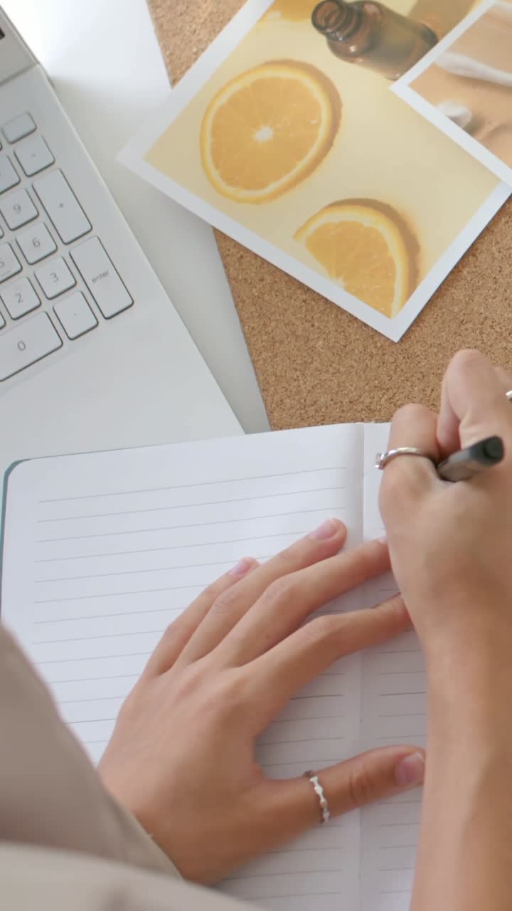 Vertical video: Writing female hands holding pen on lined pad at desk, camera panning citrus prints