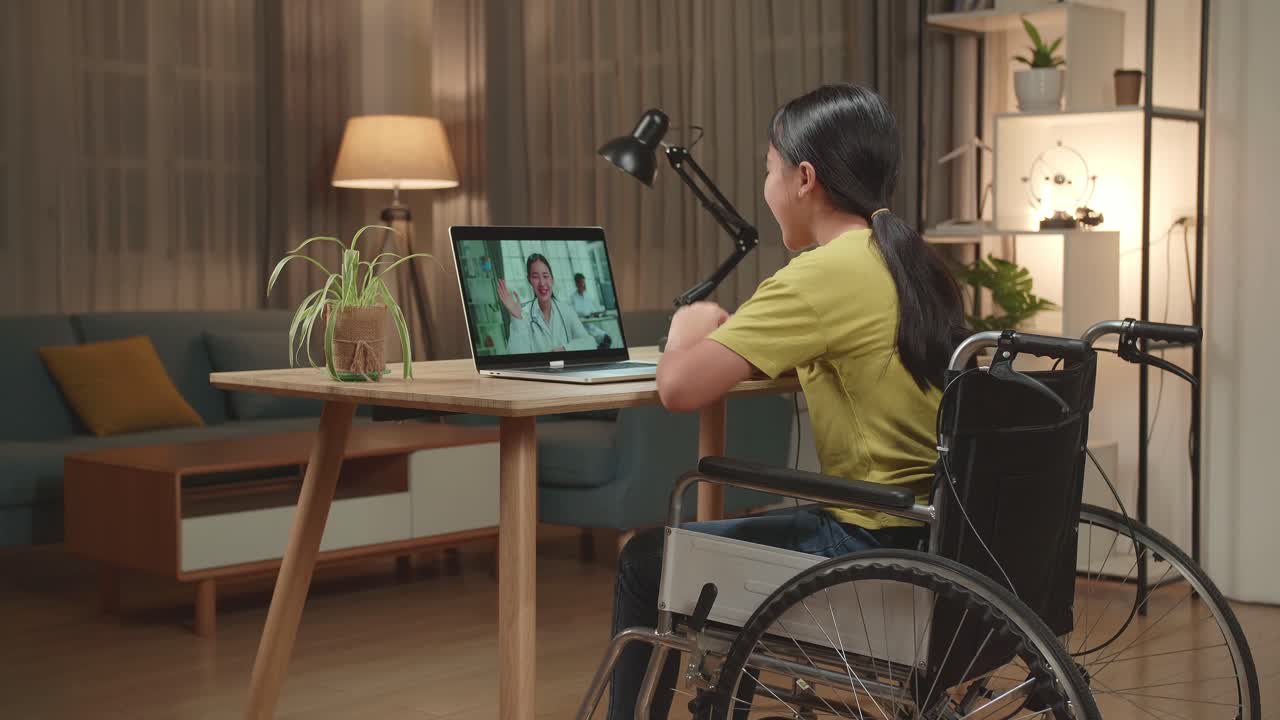 Back View Of Asian Kid Girl Sitting In A Wheelchair While Video Call With Doctor On Laptop Computer At Home