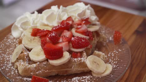 gofre belga con crema batida, fresas y plátano comido por una mujer joven con tenedor y cuchillo, postre sabroso