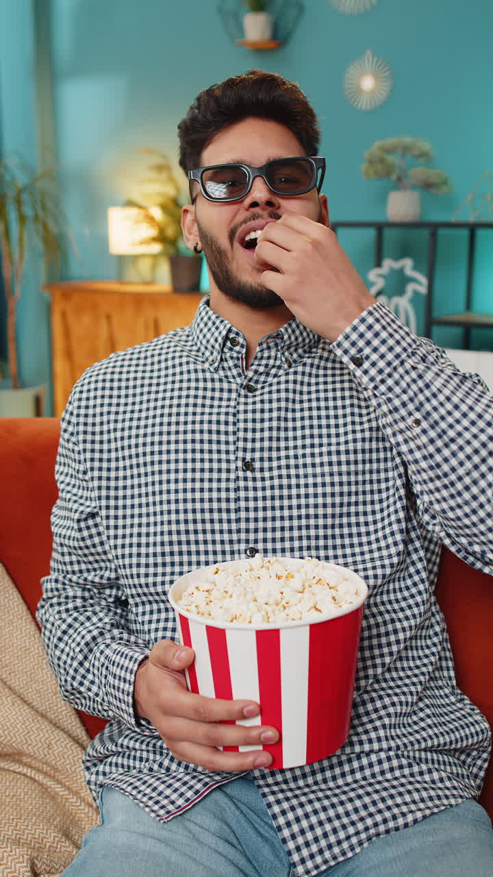 Indian man relaxing wearing 3d glasses eating popcorn and watching movie tv film at home couch