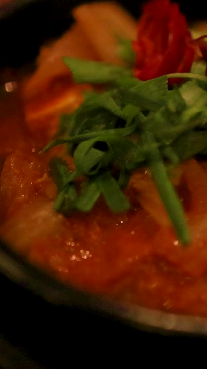 primer plano de sopa de kimchi burbujeante en un cuenco de piedra, adornada con verduras frescas, bajo una iluminación ambiente cálida