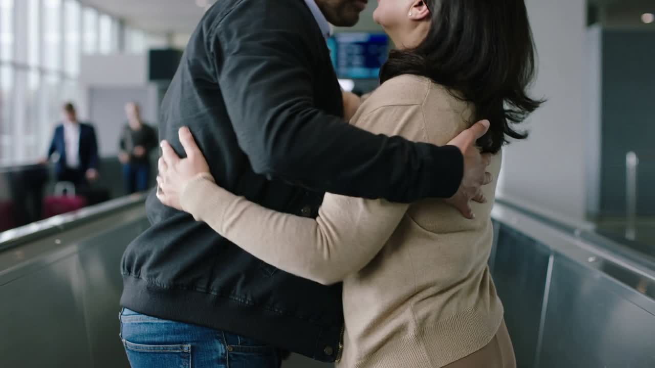 Una coppia con giacca scura e cardigan beige si abbraccia su una passerella al gate con il tabellone dei voli, ricongiungendosi.