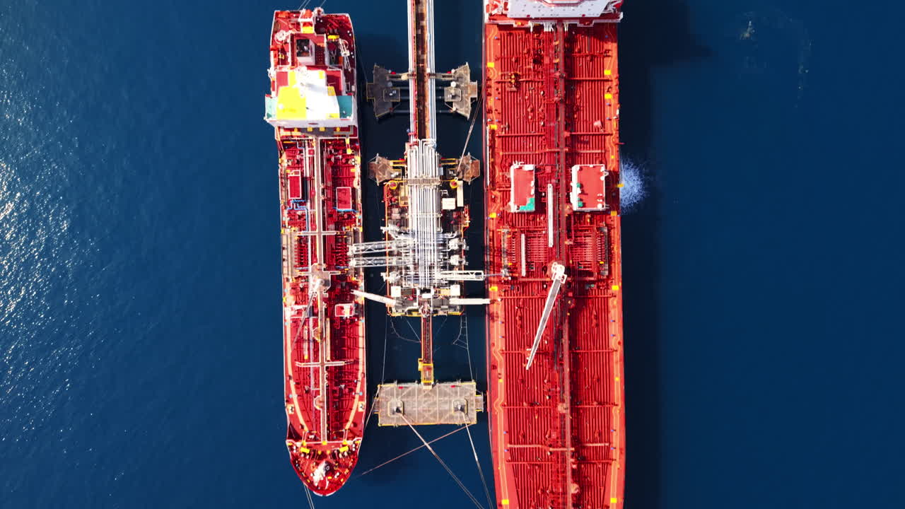 Drone shot in full top-down view moving along two red oil tankers docked at a pier over deep blue sea