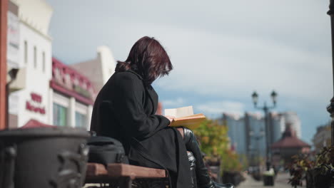 young girl in black coat reading her book outdoors, focusing on the pages while her hair blows in the wind, urban buildings, lamp poles, and passing people form a soft blurred background